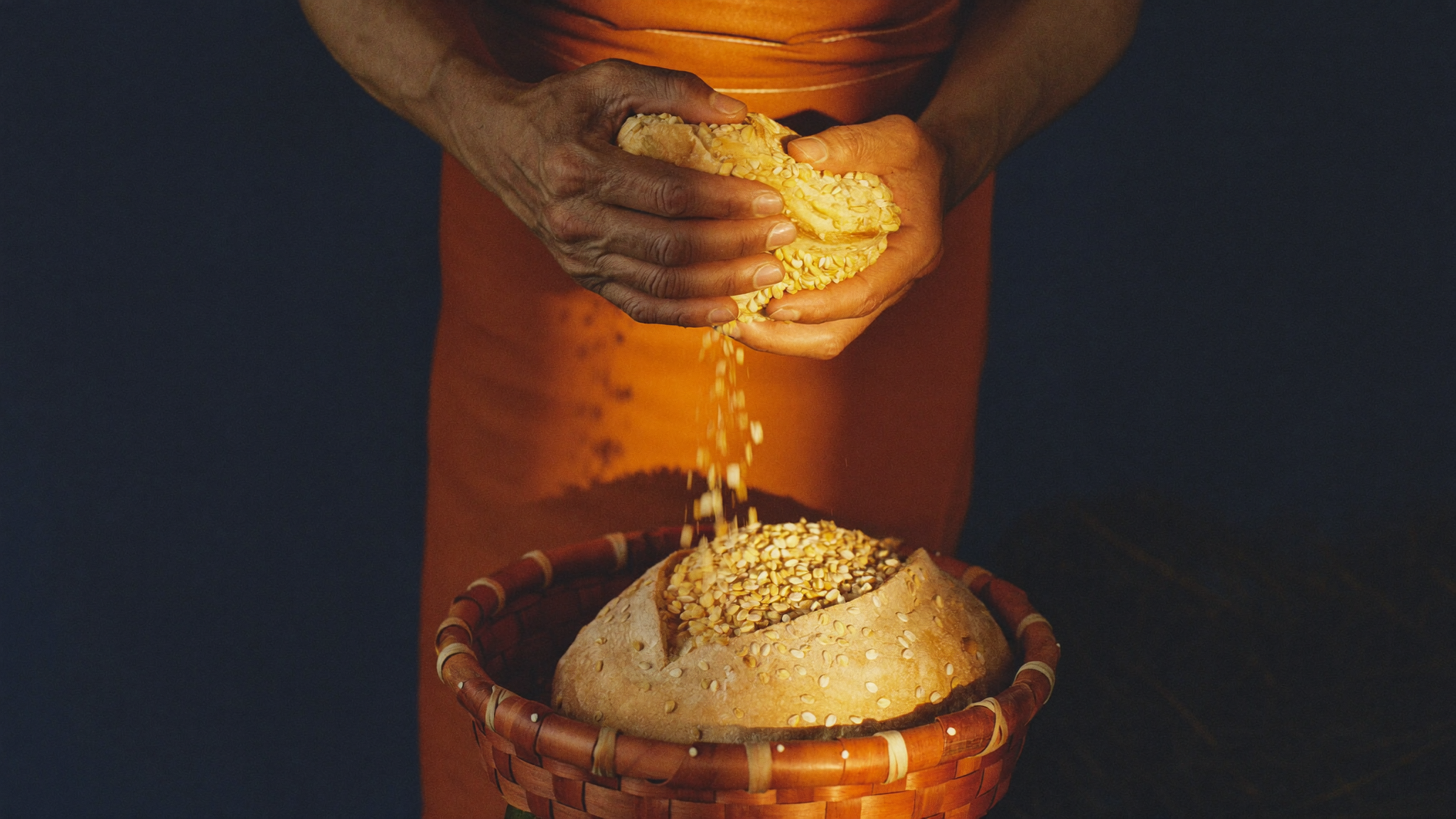 Weathered hands breaking barley bread, more loaves than the small basket could hold spilling into the light