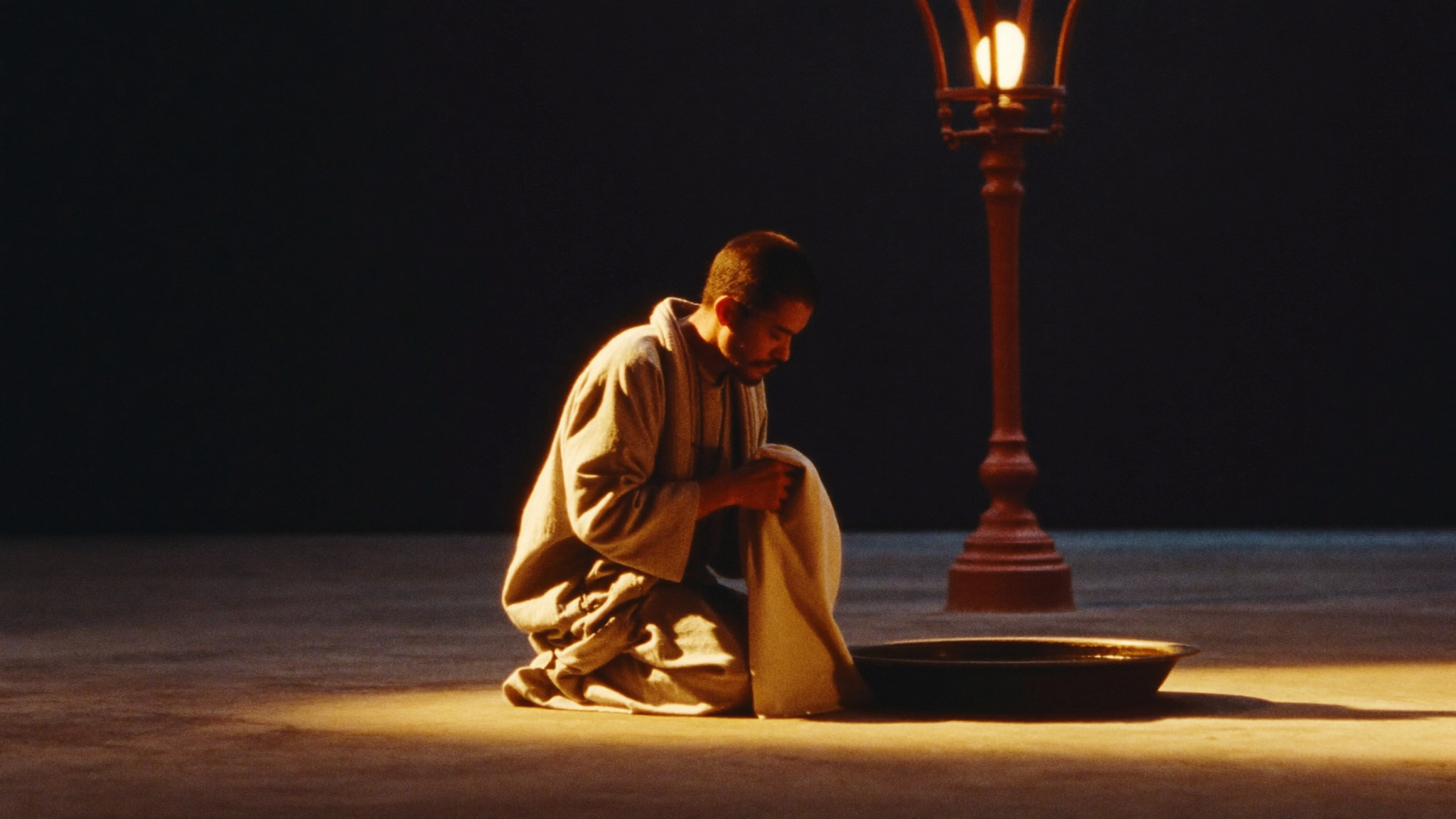 A man in plain robes kneeling on a stone floor with a towel at his waist, washing the feet of seated companions in warm golden lamplight against a dark room