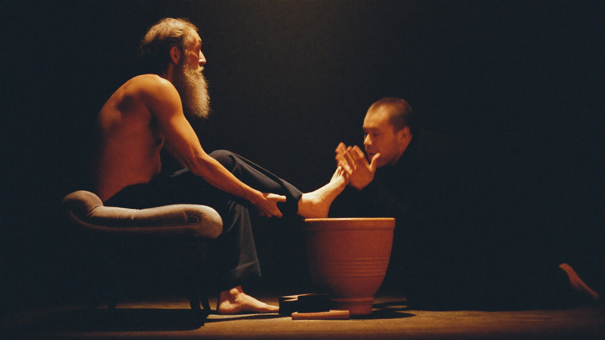An older man seated, pulling his foot back in surprise while another man kneels before him with cupped hands, both faces lit softly