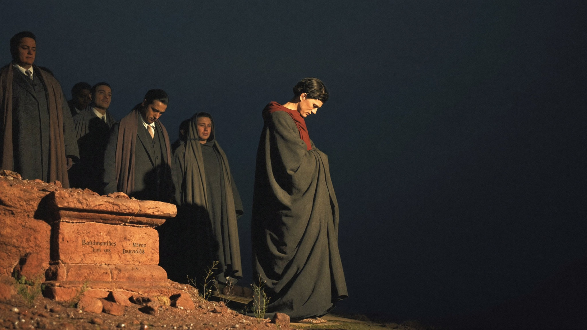 A robed man standing at the edge of a group of mourners, head bowed, tears on his face